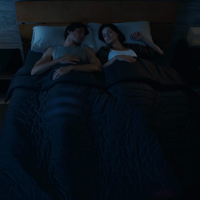 A couple asleep under one duvet, his side lit cool blue and hers warm red